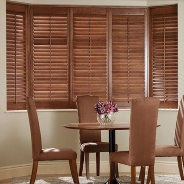Wooden horizontal blinds (bay window) closed, slats slightly filtering light; a small dining area with round table, four brown upholstered chairs and glass vase of purple flowers against neutral walls.