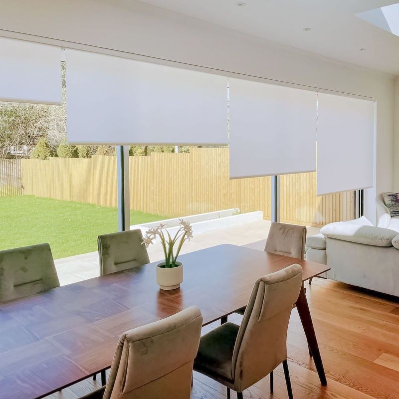White roller blinds, four adjacent panels lowered at staggered heights, partially filtering daylight over floorâ€‘toâ€‘ceiling windows; bright open dining-living area with wooden table, upholstered chairs, potted plant and lawn view.