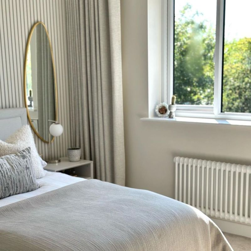 Full-length beige pleated curtains, floor-to-ceiling and drawn to the left, drape in soft folds, gently filtering daylight from an adjacent open window in a neutral, sunlit bedroom with garden view.