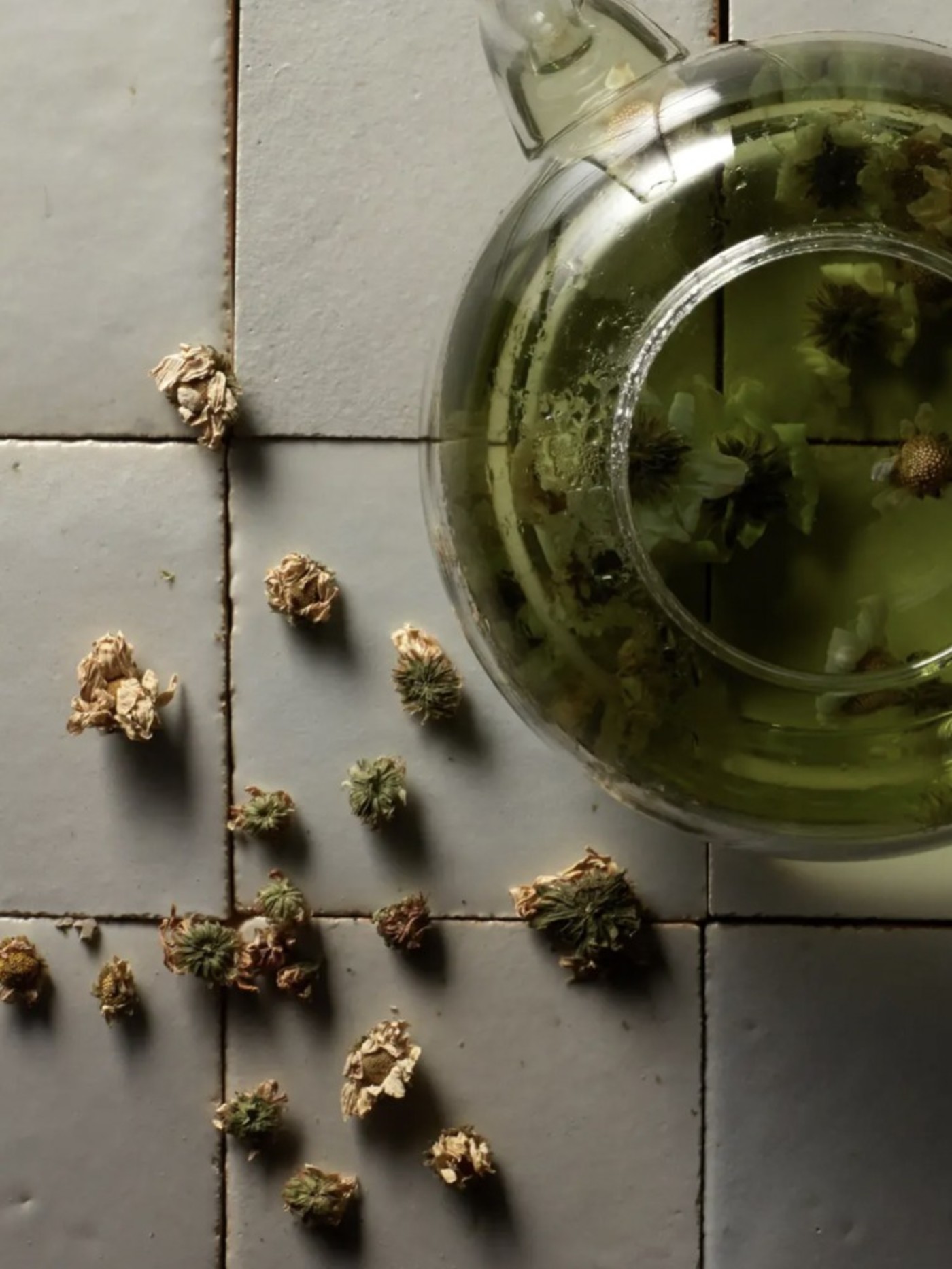 several dry flower buds next to a green glass pitcher on a tiled surface.