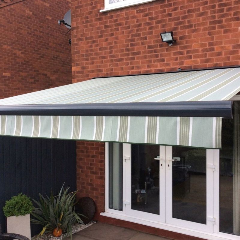 Striped retractable awning extended over a patio, pale green and white vertical stripes with a scalloped valance, shading white French doors on a red-brick house exterior with plants.