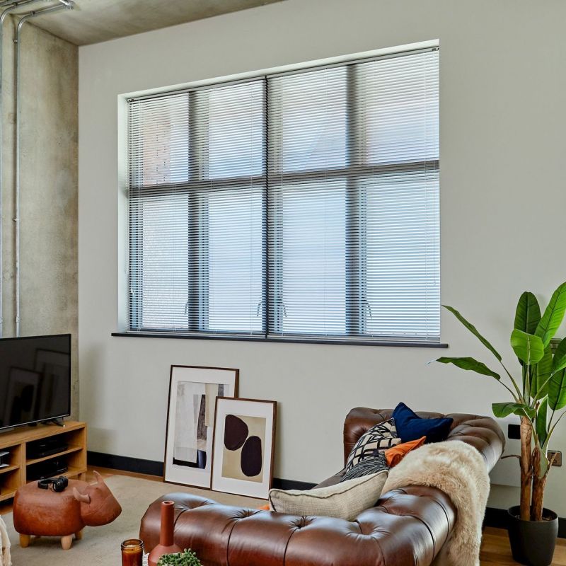 Horizontal aluminum venetian blinds covering a large industrial-style window, fully lowered and tilted closed, softly diffusing daylight into a modern living room with leather sofa, houseplant, TV and framed art.