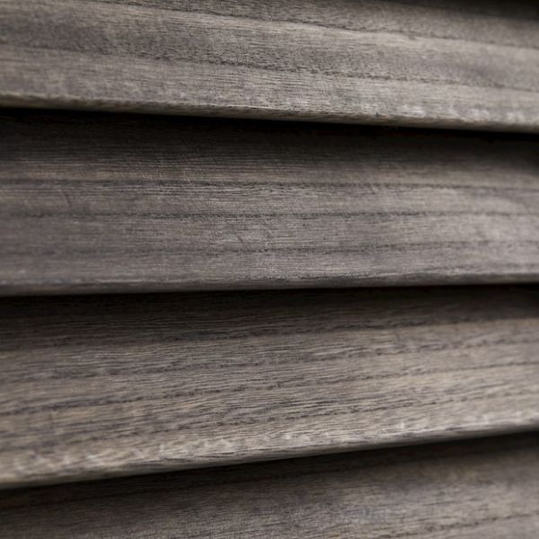Horizontal wooden blinds, narrow slatted wood shown in close-up; closed and overlapping, textured grain visible; filtering/diffusing light in a dim interior setting with neutral gray‑brown tones.