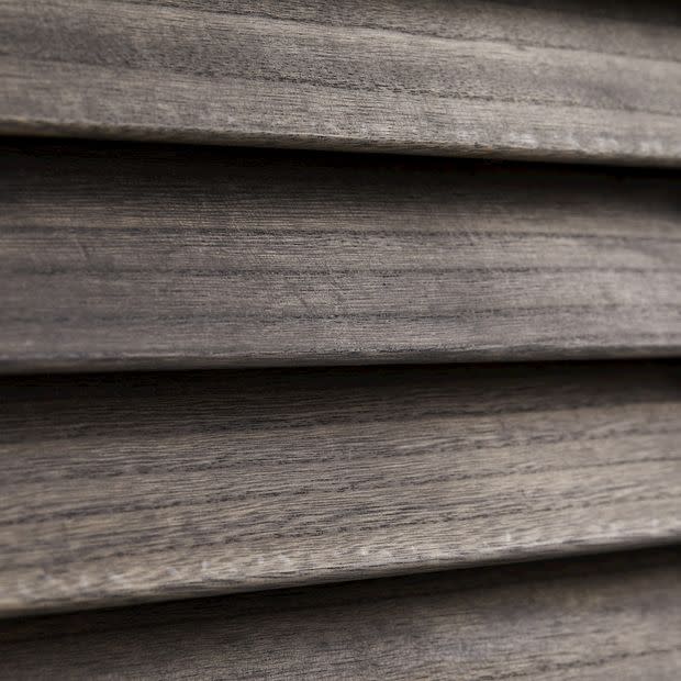 Horizontal wooden blinds, narrow slatted wood shown in close-up; closed and overlapping, textured grain visible; filtering/diffusing light in a dim interior setting with neutral grayâ€‘brown tones.