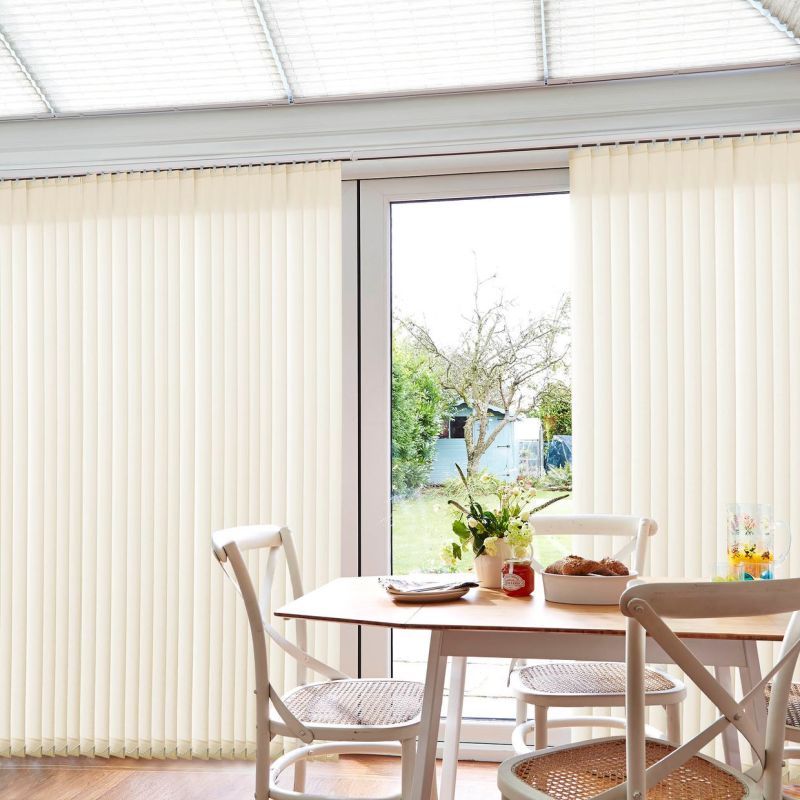 cream-colored vertical blinds cover sliding glass doors, parted at center, filtering bright daylight; small wooden dining table with white chairs, vase and bowl in sunlit dining area overlooking a garden