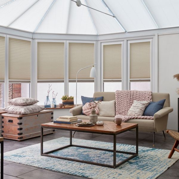 Beige pleated blinds across tall bay conservatory windows, lowered and filtering soft daylight; sunlit living area with neutral sofa draped in chunky pink throw, coffee table, chest and blue rug.