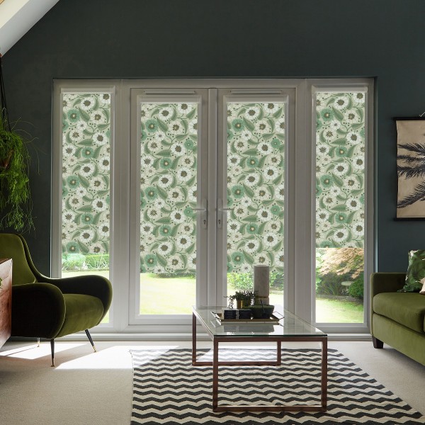Patterned floral roller blinds cover French doors and sidelights, filtering daylight in a modern living room with green velvet chairs, glass coffee table atop black-and-white chevron rug, garden visible outside.