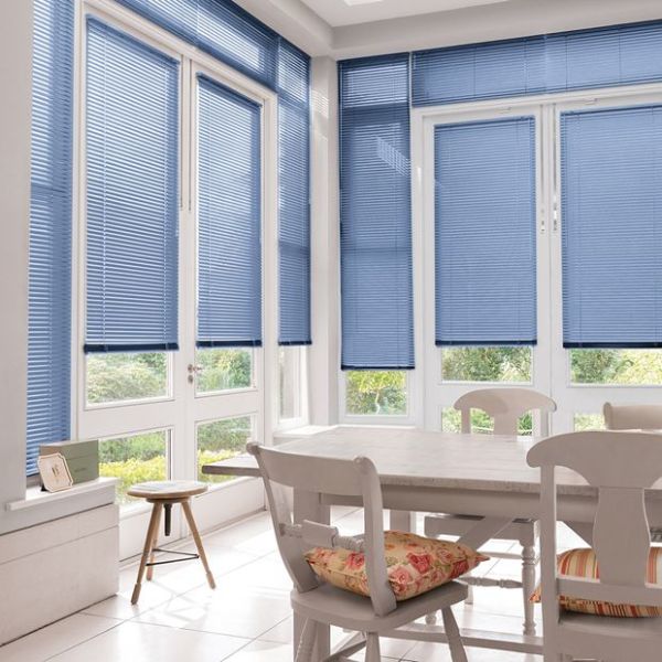 Blue horizontal Venetian blinds covering tall white-framed bay windows and door, lowered and filtering daylight; dining table and chairs in a sunlit kitchen/dining nook with cushions and garden view.