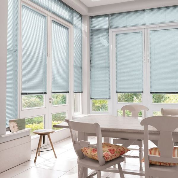 Light-blue horizontal Venetian blinds cover tall French doors and surrounding windows, lowered and filtering soft daylight; bright white dining nook with wooden table, chairs, floral cushions, tiled floor, garden beyond.