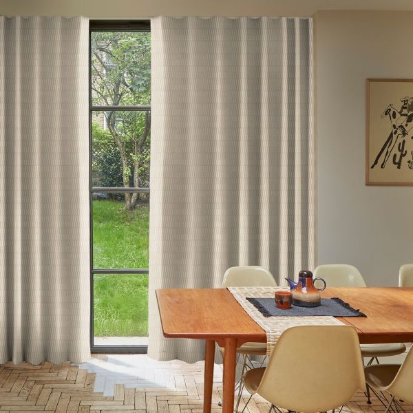 Beige textured floor-to-ceiling curtains, hung across a tall three-pane window and slightly parted, filtering soft daylight into a dining room with a wooden table, chairs, ceramic pitcher, and garden view.