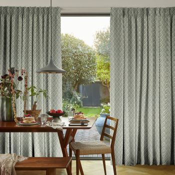 Patterned pleated floor-to-ceiling curtains, drawn apart at the center to frame the opening, revealing sunlight and a leafy backyard; wooden dining table set with breakfast, pendant light overhead.