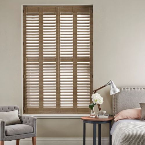Full-height wooden plantation shutters with four panels and louvers, closed with slats tilted to filter daylight; set in a neutral-toned bedroom beside a tufted armchair, bed, and bedside table lamp.
