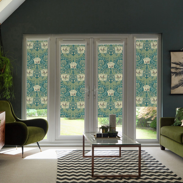Patterned floral roller blinds hang lowered over glass patio doors, filtering sunlight into a contemporary living room with olive-green armchairs, glass coffee table, zigzag rug, and houseplants.