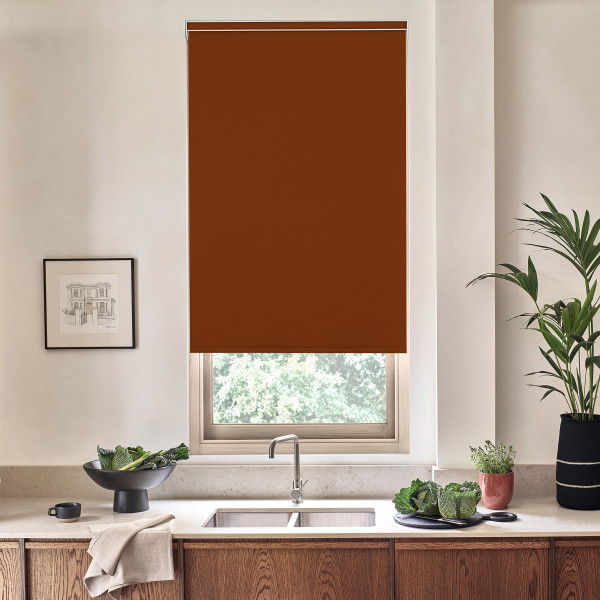 Brown roller blind covers most of a kitchen window above a stainless sink, complementing a countertop with wooden cabinets, a bowl of greens, potted plants, and a framed sketch on the wall.