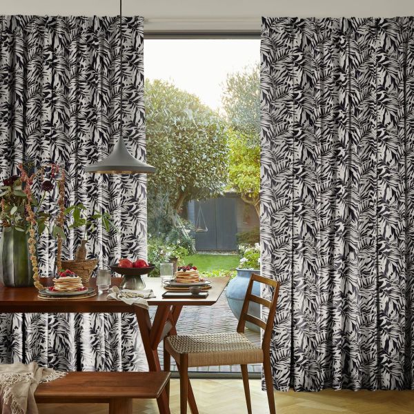 Black-and-white botanical print full-length curtains, pleated and pulled aside to frame a sliding glass door; filtering soft morning light onto a dining table set for breakfast, with a garden visible beyond.