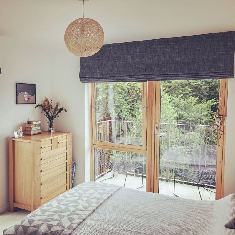 Dark-gray linen Roman blind, lowered across wooden-framed patio doors with soft folds; filtering daylight into a bright bedroom with bed, wooden dresser, ceiling light and balcony view.
