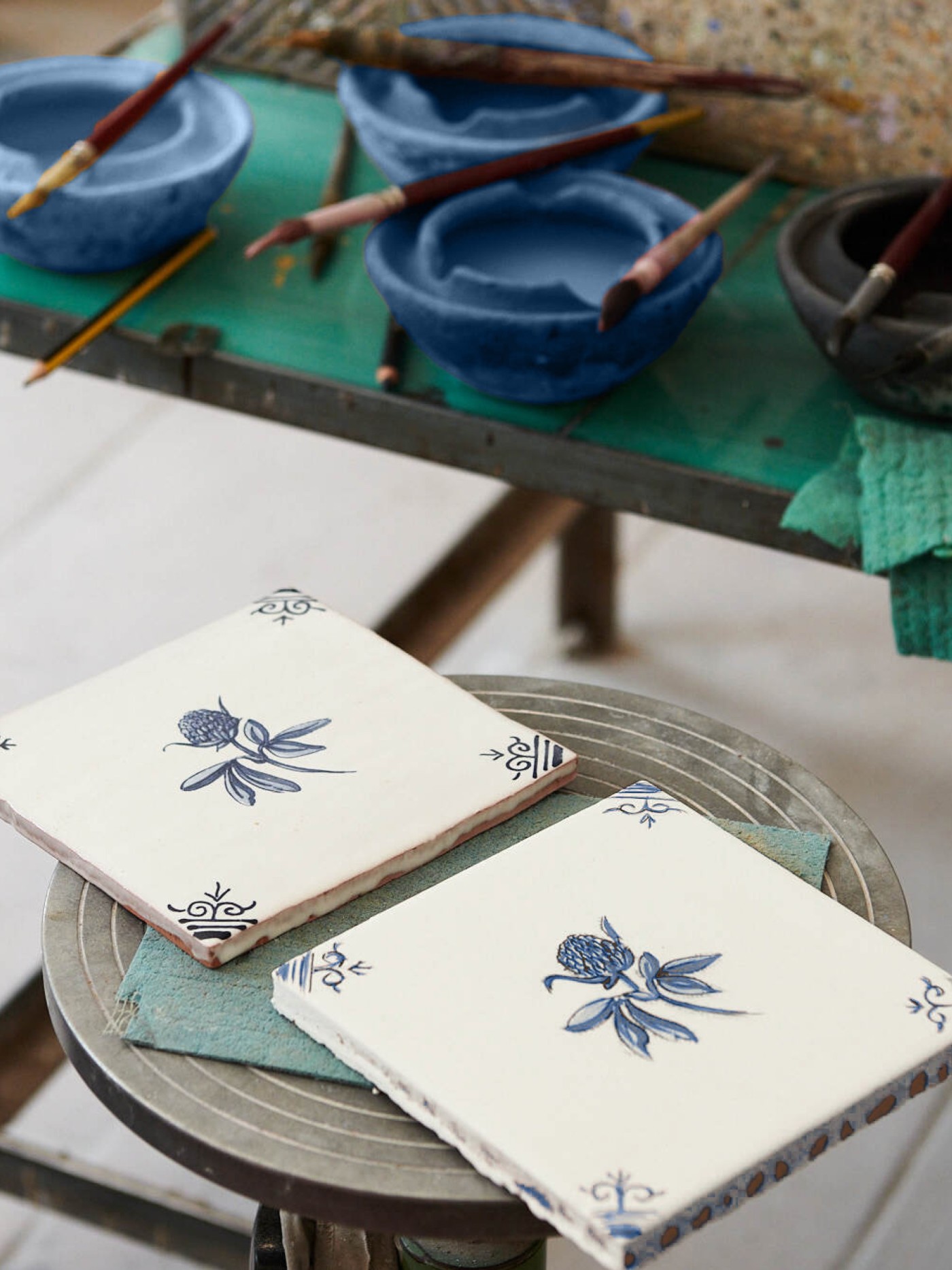 a pair of hand painted delft tiles in front in an art studio with bowls of paint and brushes in the background.