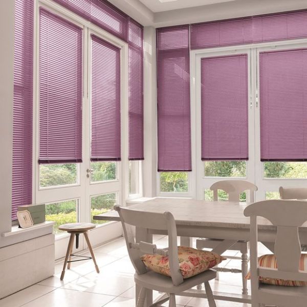 Purple horizontal venetian blinds covering tall bay windows and French doors, partially lowered and filtering soft daylight, in a bright white dining nook with table, chairs, cushions and garden view.