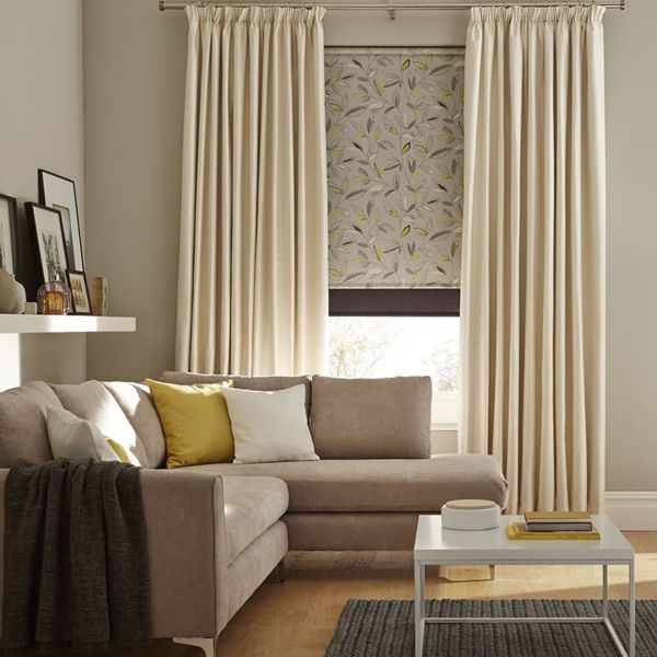Cream pleated curtains flank a patterned leaf roller blind, the blind partially lowered and filtering daylight; in a neutral living room with a beige sectional, yellow and white cushions, coffee table, shelf.