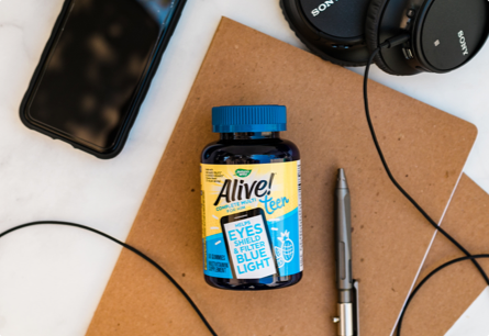 A bottle of Alive Teen Gummy Multivitamin for Him gummies laying on top of a notebook next to a cell phone, a pair of headphones, and a silver pen.