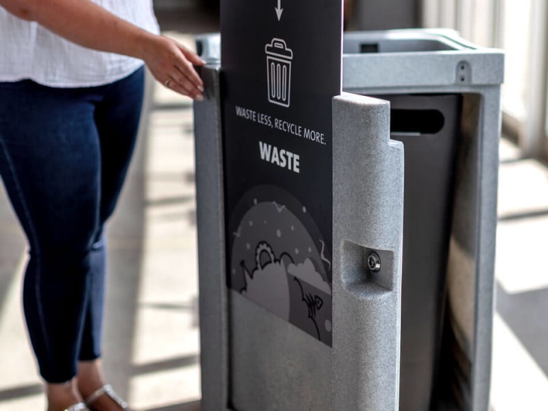 indoor grey plastic trash and recycling bin with removable lid and large body signs in the entrance of a hockey arena showing a woman servicing the bin