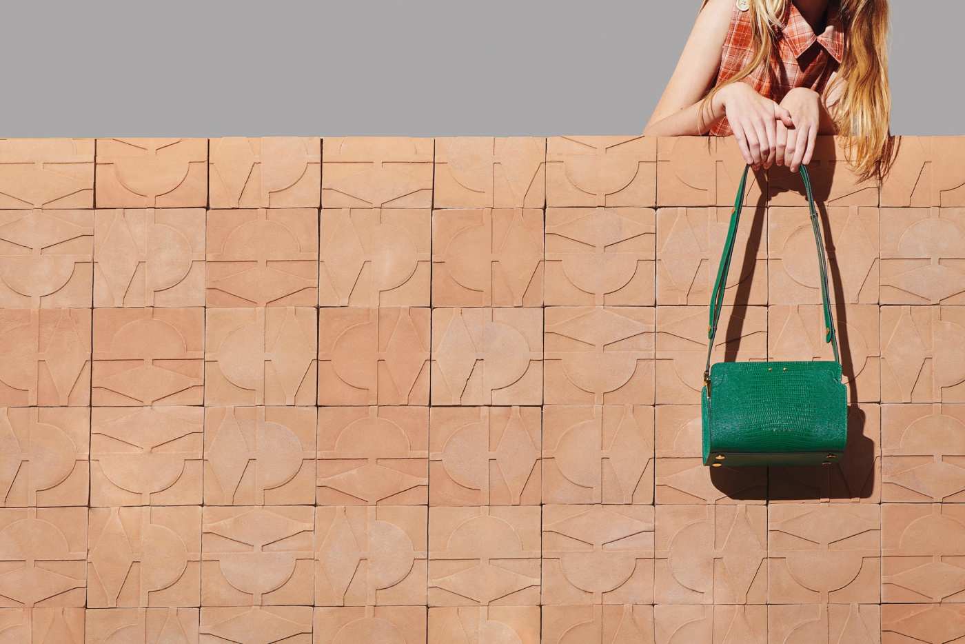 a woman with a green bag leaning against a pink tiled wall.