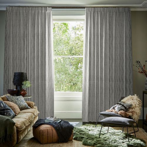 Floor-to-ceiling pleated gray drapes, slightly parted at center, filtering light; open sash window shows leafy green view; cozy living room with sofa, pouf, chair and textured rugs.