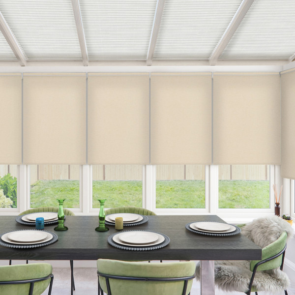 Dining table set with plates, small colored candles and green glass vases sits in a bright sunroom with beige roller blinds partly lowered, green chairs, and a grassy yard outside.