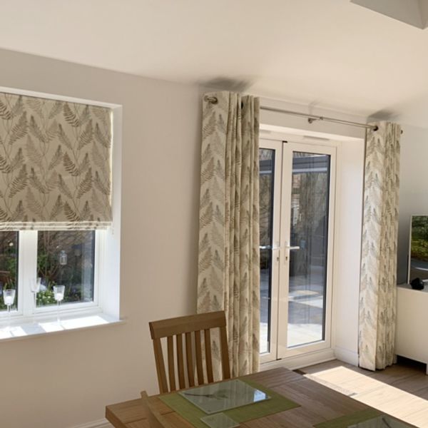 Grommet-top full-length curtains with fern print pulled to each side of French doors; matching Roman blind lowered over window, filtering sunlight; dining area with wooden table and visible patio beyond.