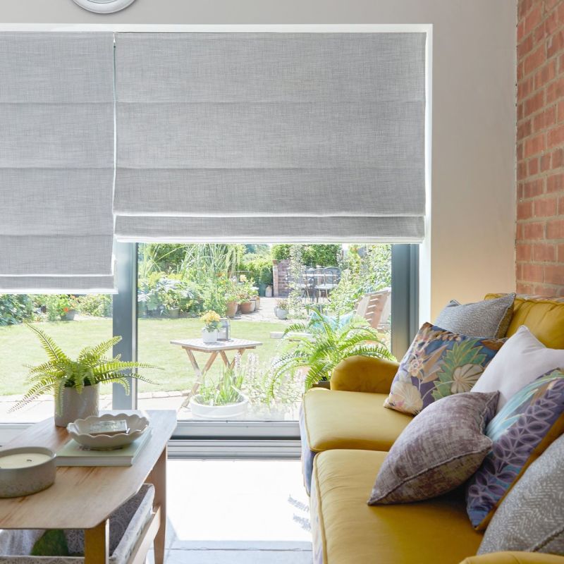 Light-gray Roman blinds, partially lowered with soft horizontal folds, filtering daylight; living room with mustard-yellow sofa, patterned cushions and coffee table; sunlit garden patio visible outside.