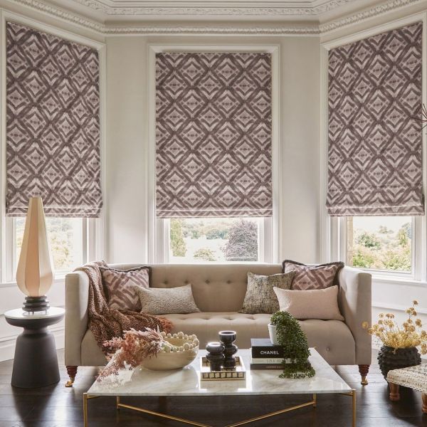 Patterned Roman shades (mauve-gray diamonds) cover three bay windows, lowered to softly filter light and partially obscure the view; neutral living room with tufted beige sofa, marble coffee table, plants.