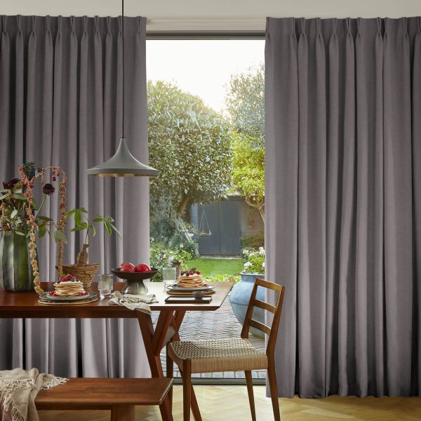 Grey pleated floor-to-ceiling curtains, slightly parted at center revealing glass door and garden; filtering soft daylight. Dining table set with pancakes and a pendant lamp in a bright dining area.