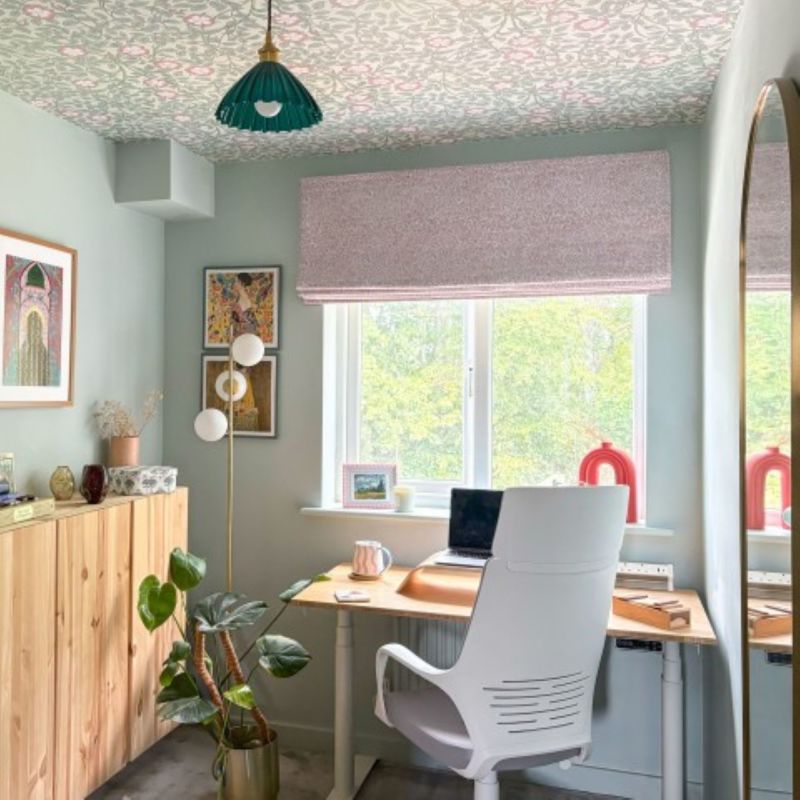 Pink patterned Roman blind over central window, lowered and filtering daylight; home office with light-wood desk, white chair, potted plants, floral wallpapered ceiling and green pendant lamp.