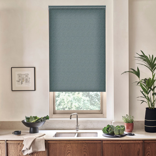 Teal cellular window shade is lowered over a kitchen window, revealing greenery through the lower pane; stainless faucet and sink rest on a stone countertop with bowl and potted plants.