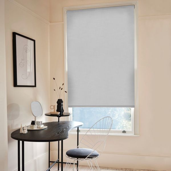 Light-gray fabric roller blind, fully lowered with a small bottom gap, filtering soft daylight over a minimalist home-office/vanity corner with pale walls, black curved desk, and a wire chair.