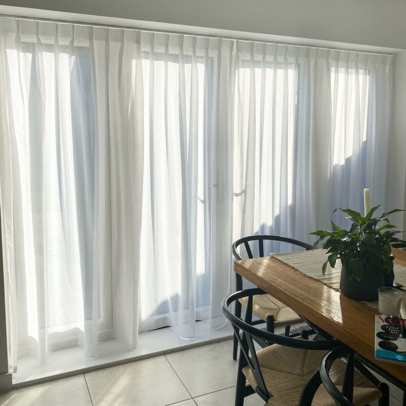 Sheer white, floor-length pleated curtains drawn closed on a wide patio door, filtering bright sunlight into a tiled dining area with a wooden table, black chairs, and a potted plant.