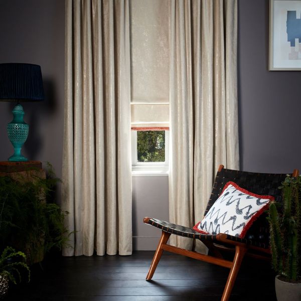 Cream full-length curtains with sheen flank a small window; a Roman blind is partially lowered, filtering daylight into a dim modern living room with dark floor and a woven lounge chair.