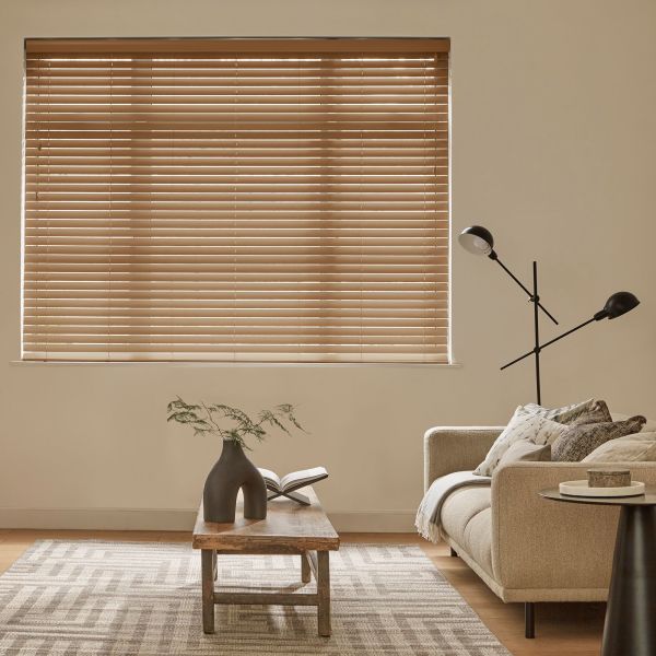 Horizontal tan Venetian blinds, fully lowered with narrow slats, filtering soft daylight through thin gaps; in a neutral-toned living room with a beige sofa, wooden coffee table, lamp, and rug.