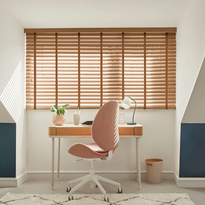 Wooden horizontal blinds covering a wide dormer window, slats tilted to filter sunlight; a compact desk with lamp, plant and pink office chair in a bright attic study.