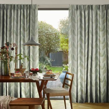 Pleated floor-length curtains with pale green fern print, drawn aside to reveal a glass door, filtering daylight into a dining nook with a wooden breakfast table and garden view.