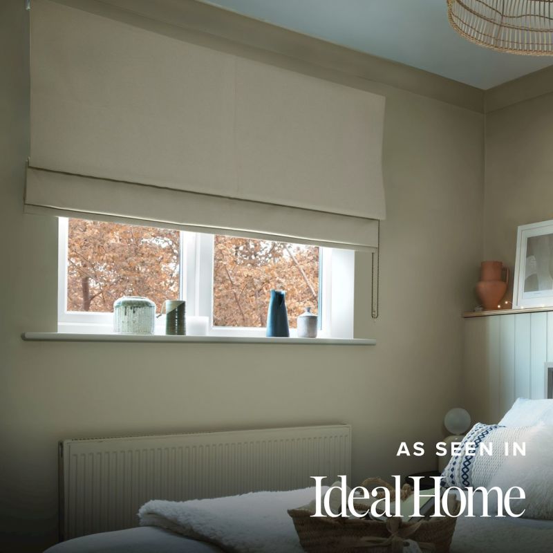 Beige Roman fabric blind, wallâ€‘mounted and partially lowered with a beaded pull, filtering soft daylight over a small bedroom window; sill holds vases, view of brown autumn trees.

AS SEEN IN IdealHome