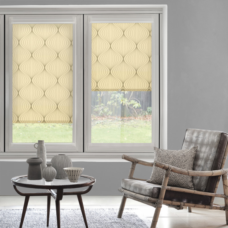 Beige roller blinds with curved pattern — left lowered, right partially lowered, filtering daylight into a gray living room with rattan armchair, cushion and round table with vases, grassy backyard view.