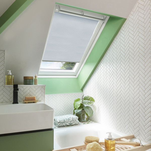 White pleated cellular skylight blind, partially lowered inside the roof window, filtering soft daylight into an attic bathroom with green trim, white herringbone tiles, sink, bathtub, plant, and bath tray.