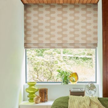 Beige wavy-patterned Roman blind lowered over the window, partly raised at the bottom; filtering daylight into a bedroom alcove with plants, glass vases on the sill and a green bedspread.