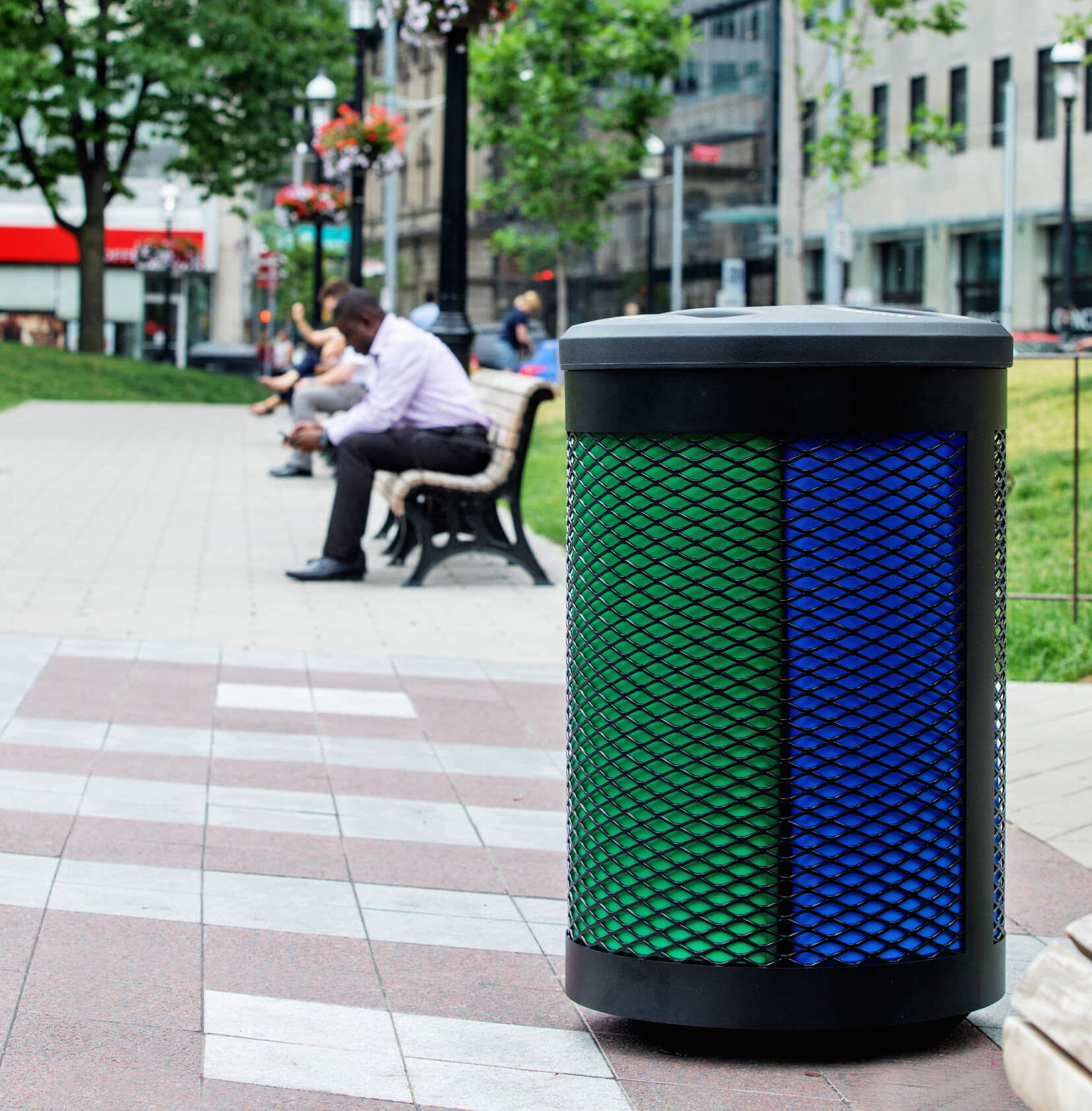 durable steel and plastic outdoor recycling and waste container with color-coded liners in a city park with a man sitting on a bench