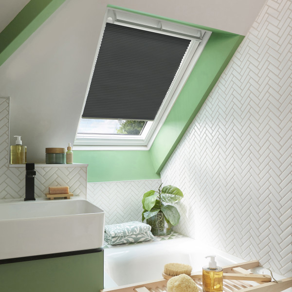 Dark gray pleated skylight blind (on an angled roof window), partially lowered and filtering daylight; bathroom with green-painted window trim, white herringbone tiles, sink, bathtub, potted plant, and toiletries.
