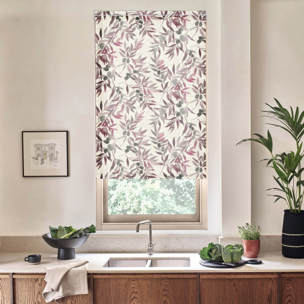 Patterned roller blind with pink and gray leaf motif covers a window above a stainless faucet and double sink, set in a bright kitchen with wooden cabinets, plants, decor.