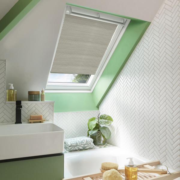Pleated beige skylight blind, lowered halfway across a roof window, filtering daylight. Set in a green-framed sloped attic bathroom with white herringbone tiles, sink, tub, plant and bath accessories.