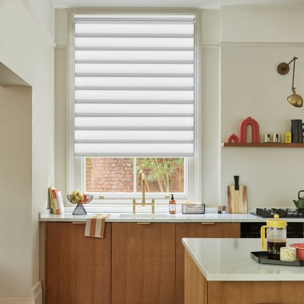 A white zebra roller blind, lowered with a small bottom gap, alternating sheer and opaque bands filtering daylight; kitchen with marble counters, oak cabinets and brass faucet, brick courtyard outside.