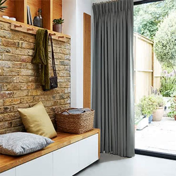 Gray floor-length pleated curtain at a sliding glass door, drawn closed, filtering daylight; a bench against exposed brick with cushions and a woven basket, patio garden visible outside.